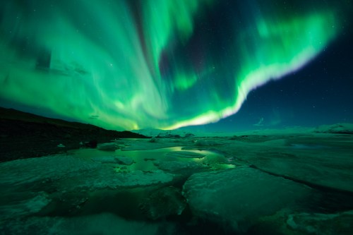 Vibrant green auroras dance across the night sky over a frozen landscape, reflecting in icy waters below.