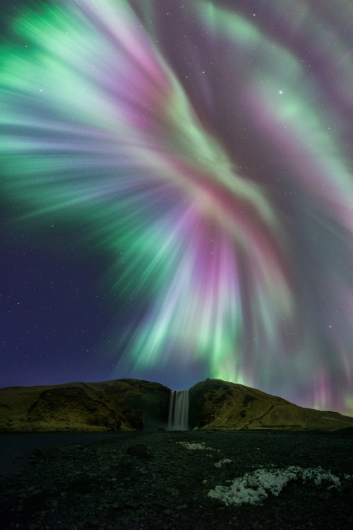 A stunning display of the Northern Lights over a waterfall, with vibrant green and purple hues illuminating the night sky.