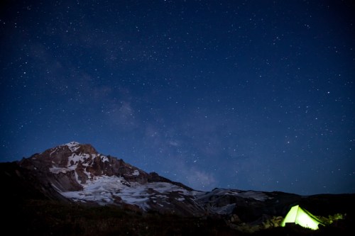 A glowing tent sits in the foreground under a starry night sky, with a mountain and glacier in the background.