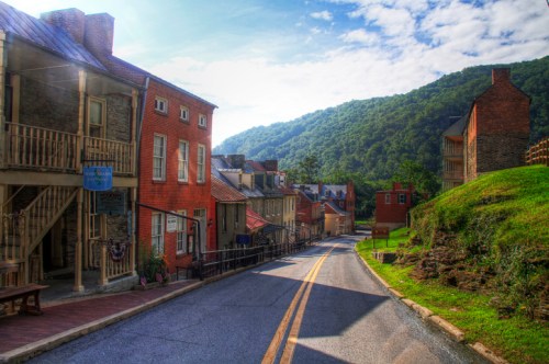 A scenic view of a quaint street lined with historic buildings, surrounded by lush green hills under a blue sky.