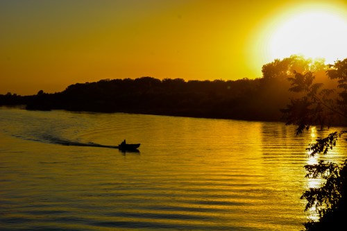 A silhouette of a boat on a shimmering river at sunset, with a golden sun setting behind trees.