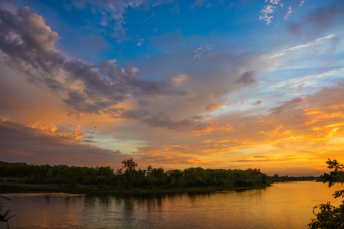 A serene river at sunset, reflecting vibrant orange and blue hues in the sky, surrounded by lush greenery.