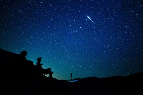 Silhouettes of two people against a starry night sky, with a bright meteor streaking across the scene.