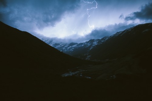 Dramatic mountain landscape under a stormy sky, with lightning illuminating the clouds and snow-capped peaks.