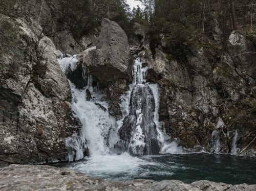 A cascading waterfall flows over rocky cliffs into a serene pool, surrounded by lush greenery and rugged terrain.