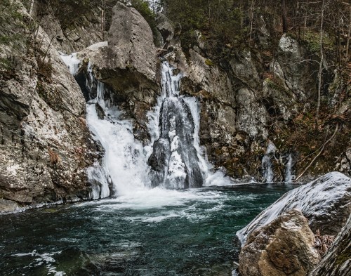 A serene waterfall cascades over rocky cliffs into a clear, turquoise pool surrounded by lush forest.