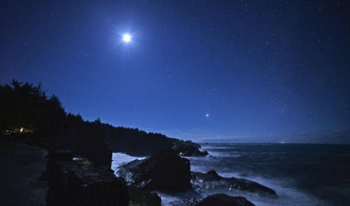 A serene night scene of a rocky coastline under a starry sky, illuminated by a bright moon and distant stars.