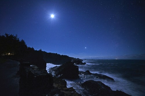 A serene night scene of rocky coastline under a starry sky, with a bright moon and planet shining above the ocean.
