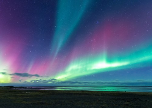 Vibrant auroras dance across a night sky, reflecting on calm waters with a serene landscape below.