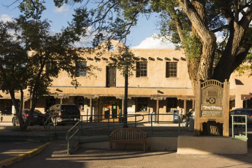A historic adobe building with a sign, surrounded by trees and parked cars, in a sunny plaza setting.