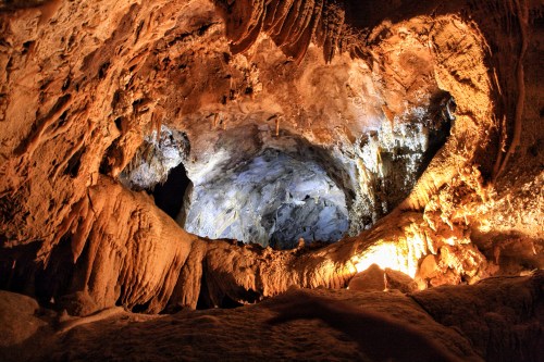 A view inside a cave, showcasing stalactites and stalagmites illuminated by soft blue and orange lighting.