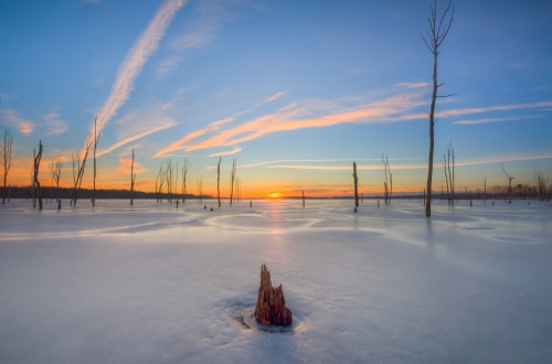 A serene winter landscape at sunset, featuring a frozen lake, silhouetted trees, and colorful sky streaks.