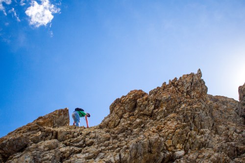 A climber ascends a rocky mountain under a bright blue sky with a few clouds.