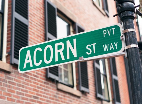 Green street sign reading "ACORN ST WAY" mounted on a black pole, with a brick building in the background.