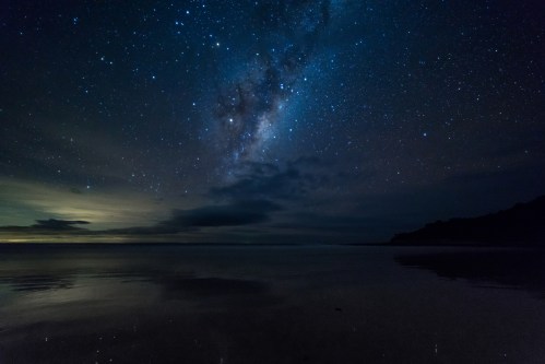 A serene night sky filled with stars and the Milky Way, reflecting on calm waters at the beach.