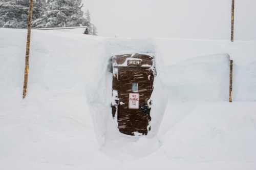A wooden restroom door partially buried in snow, surrounded by tall snowdrifts and a "No Parking" sign.