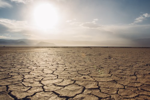 A dry, cracked earth landscape under a bright sun, with distant mountains and a hazy sky in the background.