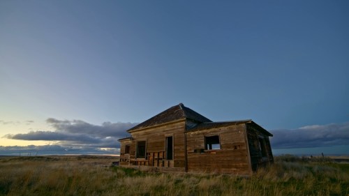 Abandoned wooden house in a grassy field under a blue sky with clouds at dusk.