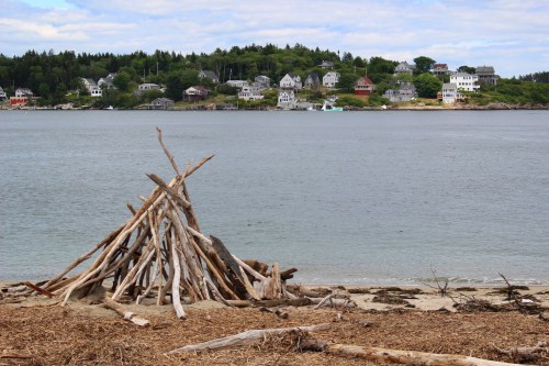 A beach scene featuring a driftwood structure in the foreground and a coastal village in the background under a cloudy sky.
