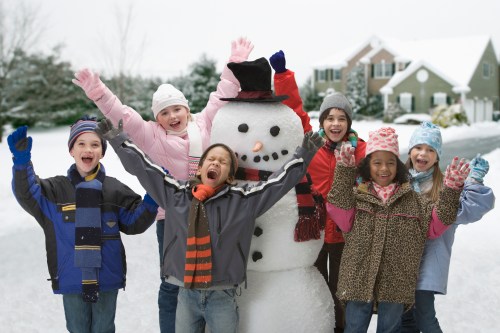 A group of joyful children in winter clothing cheer around a snowman in a snowy neighborhood.