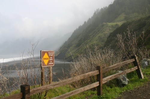 A foggy coastal scene with a warning sign about danger, surrounded by lush green hills and a wooden fence.