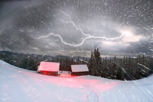 Two cabins in a snowy landscape under a stormy sky with lightning and heavy snowfall.