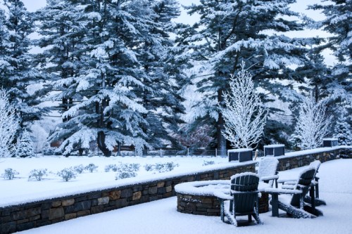A snowy landscape featuring evergreen trees, a stone wall, and a patio with chairs covered in snow.