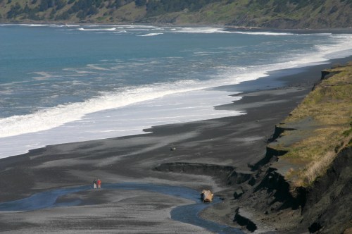 Two people walk along a black sand beach, with waves lapping at the shore and lush green hills in the background.