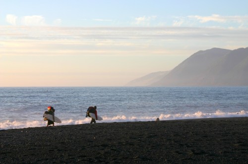 Two surfers walk along a black sand beach, carrying their boards, with mountains in the background and gentle waves.