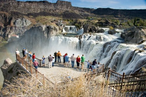 A group of people stands on a viewing platform overlooking a large waterfall surrounded by rocky terrain.