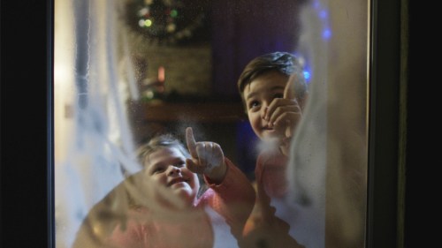 Two children smile and point at a frosted window, with festive decorations visible in the background.