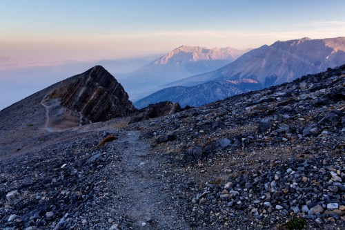 A rocky mountain trail leads through rugged terrain, with distant peaks under a soft, hazy sky at dawn.
