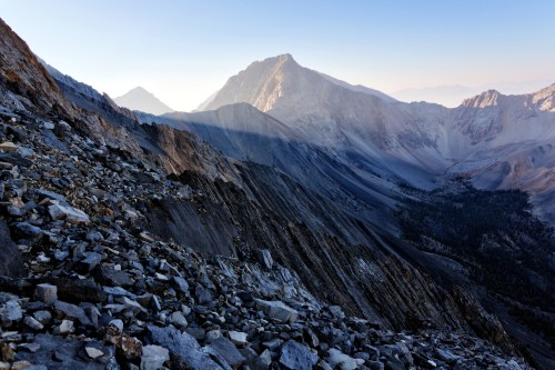 A rugged mountain landscape with rocky slopes and distant peaks under a clear sky at dawn.