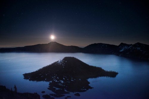 A serene night scene of a lake with a small island, illuminated by moonlight and surrounded by mountains under a starry sky.