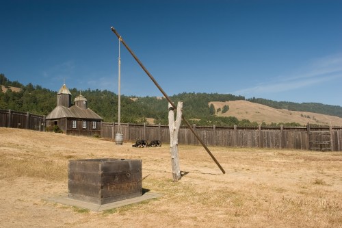 A wooden well with a long arm, set against a backdrop of a historic building and rolling hills under a clear blue sky.