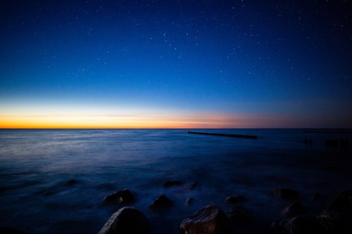 A serene seascape at dusk, featuring calm waters, rocky shore, and a starry sky transitioning from blue to orange.