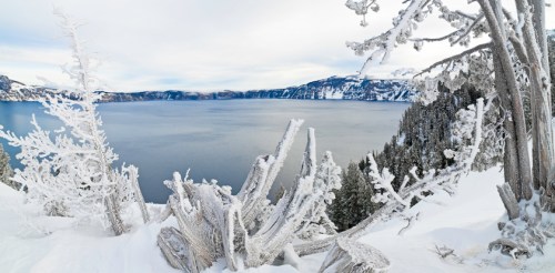 A snowy landscape overlooking a calm lake, surrounded by frosted trees and mountains under a cloudy sky.