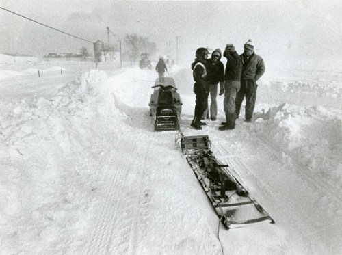 Four people stand near a snowmobile on a snowy road, surrounded by deep snow and a foggy winter landscape.
