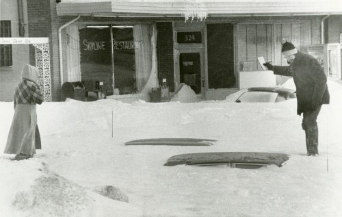 A child in winter clothing stands in deep snow while an adult gestures from a snow-covered street. A restaurant is visible behind.