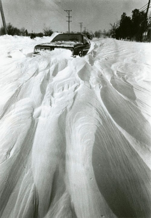 A car partially buried in deep snow on a deserted road, with snowdrifts creating wavy patterns around it.