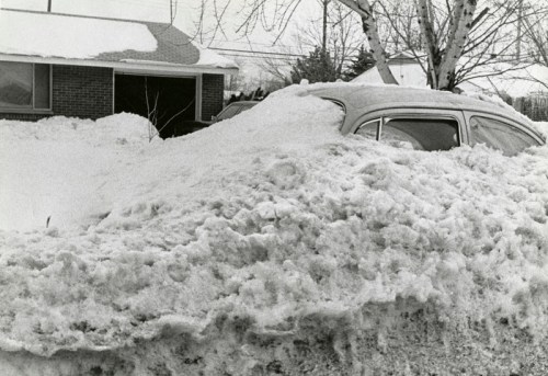A car almost completely buried in snow, with a house and bare trees in the background on a winter day.