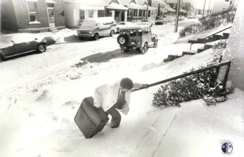 A person struggles up snowy steps with a suitcase, surrounded by snow-covered houses and parked cars.