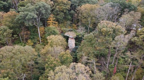 Aerial view of a tall rock formation surrounded by dense, colorful autumn foliage in a forested area.