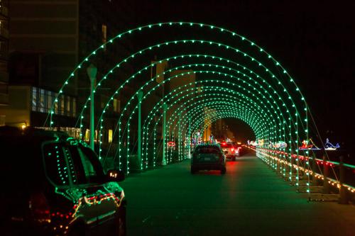 A nighttime view of a lighted archway over a road, with cars driving through and colorful decorations in the background.