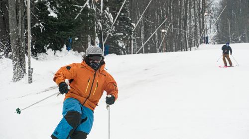 A skier in an orange jacket and blue pants carves down a snowy slope, with trees and another skier in the background.
