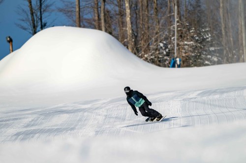 A snowboarder glides down a snowy slope with trees in the background and a snow-covered hill in the distance.