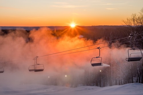 A ski lift silhouetted against a vibrant sunset, with mist rising over a snowy landscape.