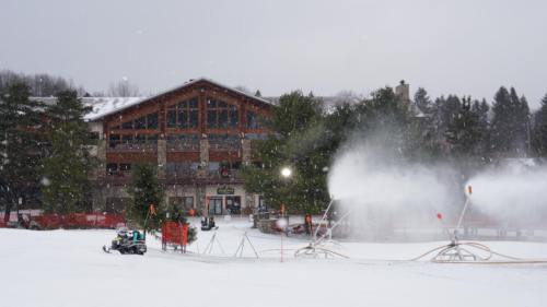 A snowy landscape with a lodge in the background and snowmaking equipment in the foreground.
