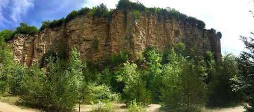 A panoramic view of a rocky cliff surrounded by lush green trees under a bright blue sky.