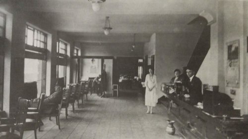 A vintage interior of a waiting room with chairs, a reception desk, and three people standing near the counter.